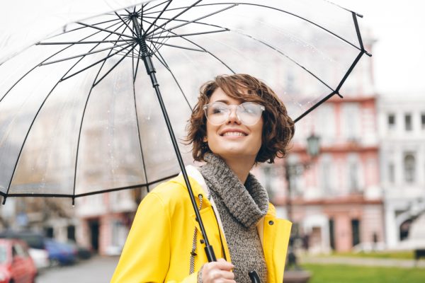 Amazing portrait of happy woman in yellow raincoat walking in city under transparent umbrella, during cold rainy day